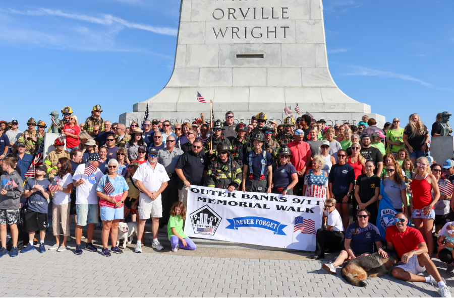 A large group of people stand in front of the Wright Brothers Monument holding a sign that reads," Outer Banks 9/11 Memorial Walk | Never Forget"
