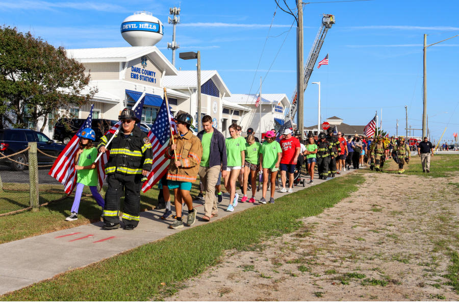 Image of people walking in front of the EMS station carrying American flags. Some wear firefighter uniforms.