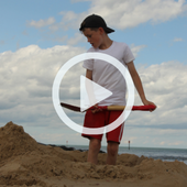 A boy digs a hole in the sand on the beach. A play button signifies that a video will play when clicked.