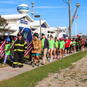 A line of people in patriotic clothing walk along the sidewalk in front of the Dare County EMS Station 1 and Kill Devil Hills water tower.