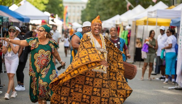 A Black man and woman in vibrant, flowing attire dance down Fayetteville Street between artist booths.