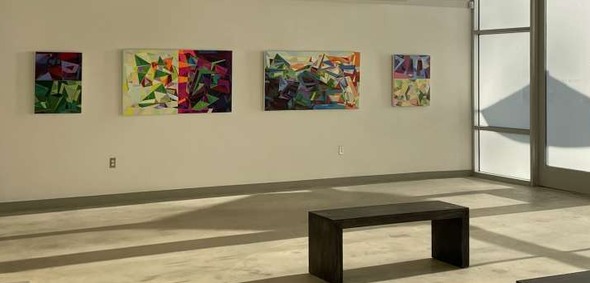 White-walled gallery space with large colorful, abstract paintings with sunlight streaming in from large windows. 