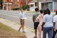 A tour guide leads a group of listeners and points out historic houses in the background.