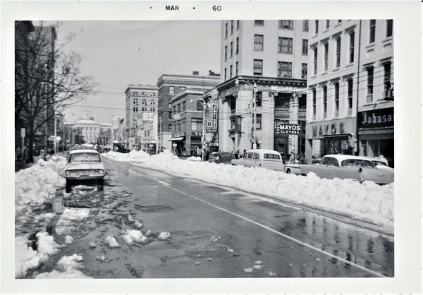 Fayetteville Street with snow piled on the sides and middle of the road. Cars are parked and the State Capitol is visible in the distance. 