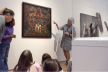 A group of students looks at a painting as a tour guide points out details from it.] 