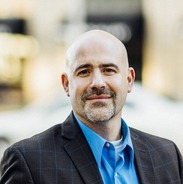 Waist-up image of Dr. Shawn Oban with a bald head, closed smile, and goatee, wearing a blue collared shirt and dark grey suit jacket