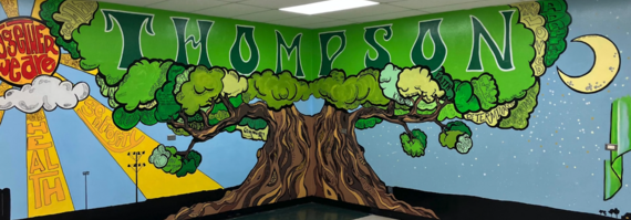 Colorfully painted mural of a huge tree with a sun and a half moon on two walls in the cafeteria at Thompson Public School