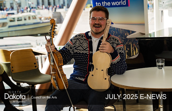Laughing Gus Holley holding two special commemorative fiddles, wearing a Norwegian sweater, sitting on the inner deck of a cruise ship in Oslo, Norway