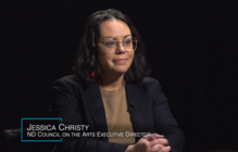 NDCA Executive Director Jess Christy, sitting behind a desk, on a black background
