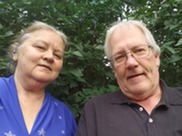 half-smiling, grey-haired, caucasian, Connie and Rick Whittier with green leaves behind them