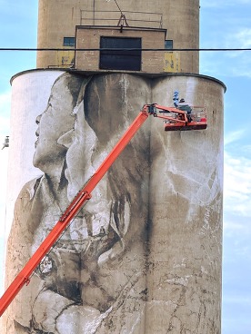 Austrailian artist Guido van Helten on a lift, painting a woman and child on one side of a half-dozen grain silos that will because ND's largest mural