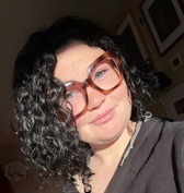 head & shoulders of Zoe Makin with curly, shoulder-length dark hair, thick-rimmed brown glasses, blue eyes, closed smile, wearing a sweatshirt