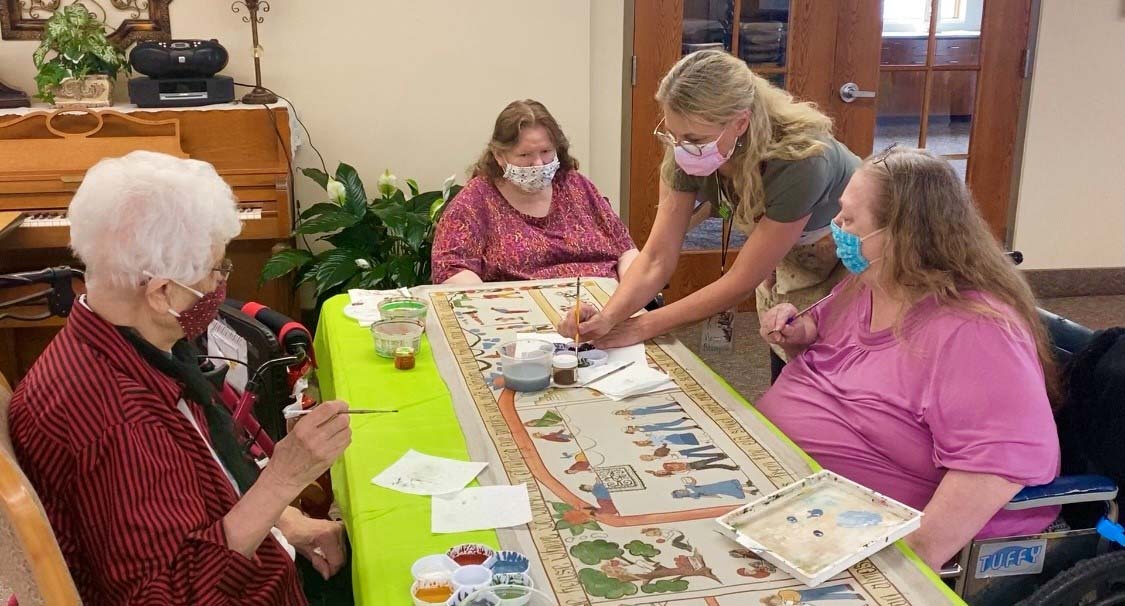 wearing masks, folk artist Pieper Bloomquist engaged elderly residents in painting on one canvas at Maryhill Manor in Enderlin, ND