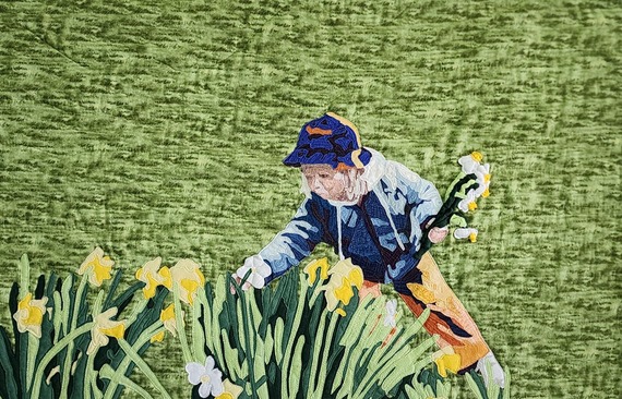 Art quilt “Nanna’s Daffodils” shows a figure in blue and orange picking daffodils in a yellow-white field with a soft green fabric background.