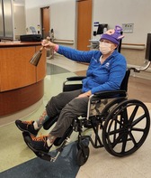 Jane Gaffrey, in a blue shirt and pink beanie, rings a bell from a wheelchair in a hospital, smiling behind a mask.