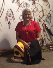 A woman in a red shirt and a colorful skirt kneels in a room decorated with handmade spiritual and artistic objects on the wall.