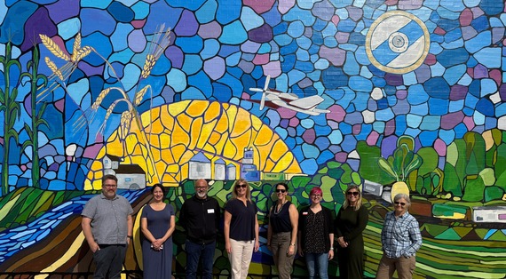 NDCA Board stands before a vibrant mosaic mural depicting a rural landscape with a river, hills, silos, and a plane in a colorful sky.