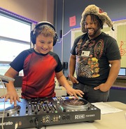 A boy with headphones smiles while learning to use a RANE PERFORMER DJ controller, as a man in a fur hat smiles behind him.