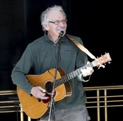 An older man with glasses and gray hair, smiling and singing into a microphone while playing an acoustic guitar.