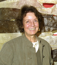 A smiling woman with brown hair in a green jacket standing in front of an art installation made of organic material.