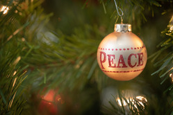 Golden ornament that reads PEACE, hangs from the branches of an evergreen Christmas tree with lights