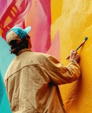 Artist working on a large, vibrant wall mural with bright yellow, pink, and teal paint.