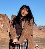 Native American woman with a toothy smile, long black hair, standing in front of canyons & blue sky, wearing a brown coat & dark brown undershirt