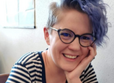 Smiling person with blue-tinted hair and glasses, wearing a striped shirt. A tattoo on their arm reads "Nevertheless, she"
