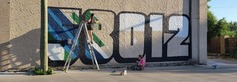 Fargo artist Lauren Starling standing on a ladder, painting a mural outside showing the 58012 zip code of Castleton, ND