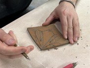 A square piece of clay that is being carved by 2 hands and a carving tool
