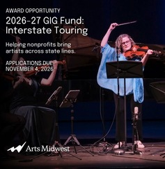 A violinist in a blue top raises their bow dramatically while performing on a darkened stage