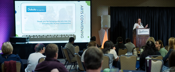 NDCA Executive Director Jess Christy standing on stage, speaking in front of a 50-60 people with a large screen on her right