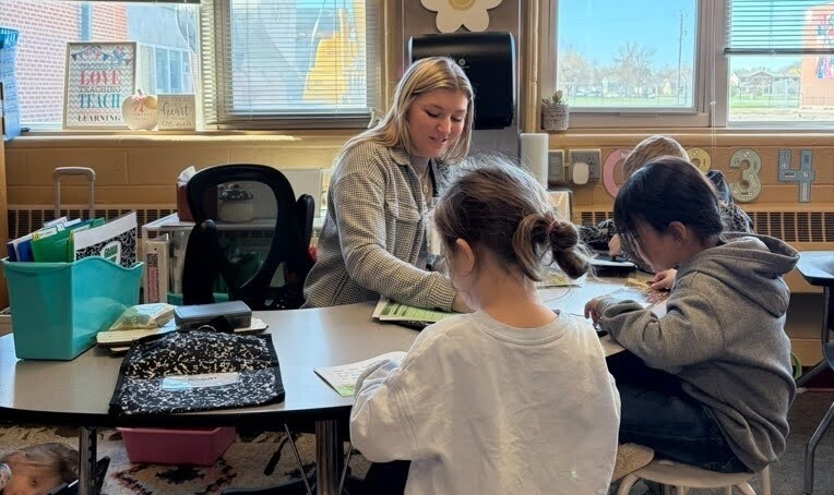 Teacher sits at table with young students learning.