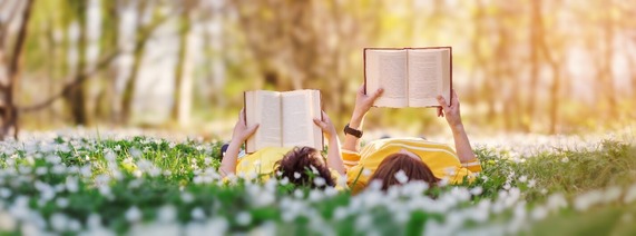 two people laying on grass reading books. Trees in background