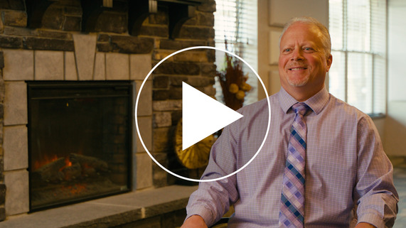 Man sitting smiling at camera with a play icon overlayed on top of the image.
