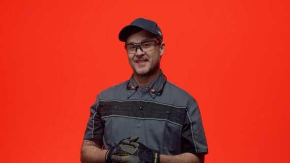 Man in work clothes, gloves, safety glasses, and hat speaking to camera against red background.