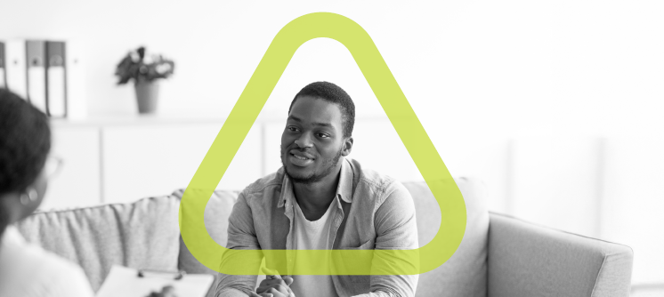 Black and white photo of man sitting speaking to a person in the left corner. The green GHS triangle surrounds him.