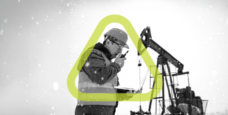 Oil field worker wearing a hard hat and safety vest uses a radio while holding a laptop near an oil pump in snowy conditions
