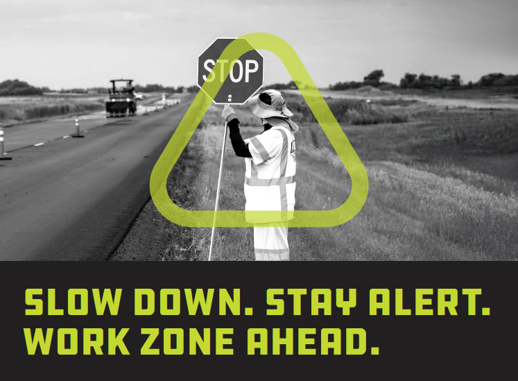 Construction sign holder standing on side of road wearing bucket hat and hi-vis gear inside the Get Home Safe triangle.