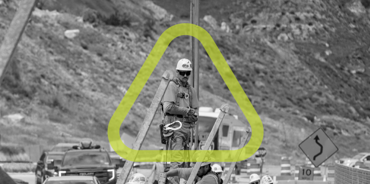 Construction worker wearing safety harness, hard hat, and sunglasses standing on a piece of equipment while cars drive by.