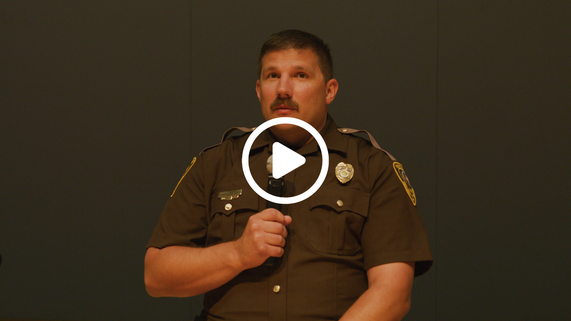 Man in a brown state trooper uniform holds a microphone while speaking to the audience with a play symbol overlaid.