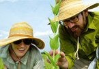 Man and woman with wide-brimmed hats in field, looking at a plant with smiles