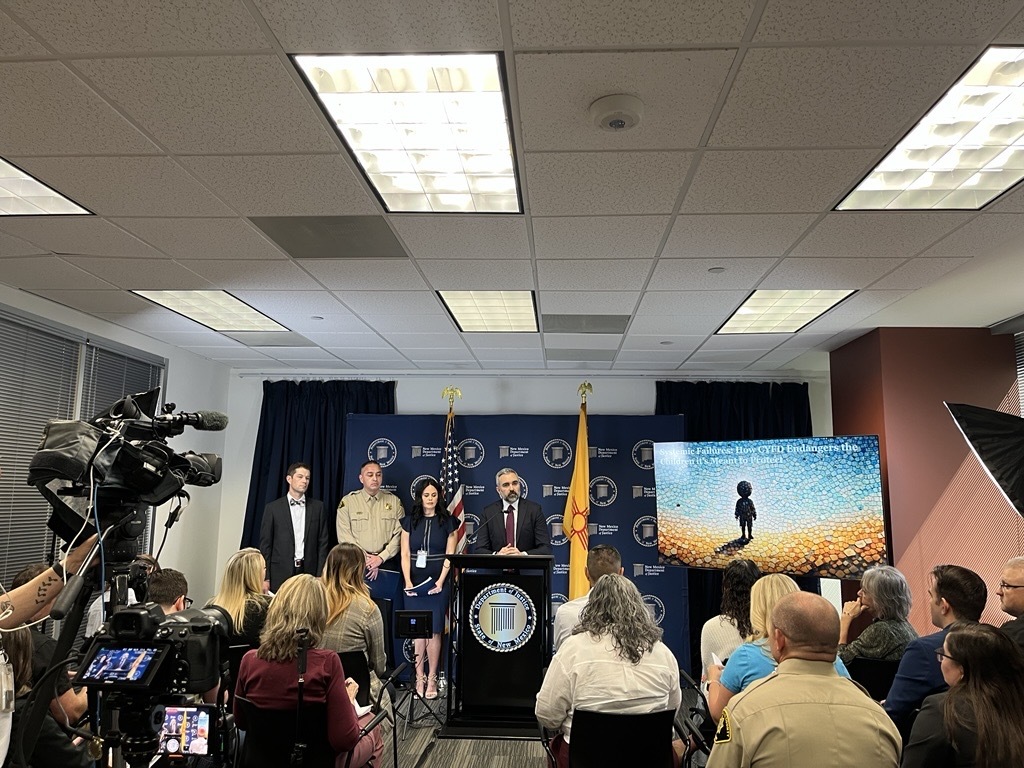Press conference with speakers at a podium, audience seated, and a screen displaying the CYFD investigative report cover.