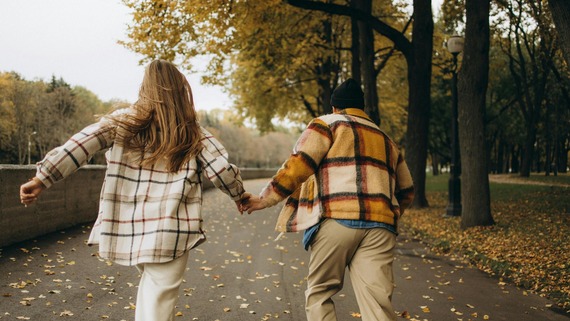 a couple wearing plaid flannels holds hands, running along a path with autumn leaves
