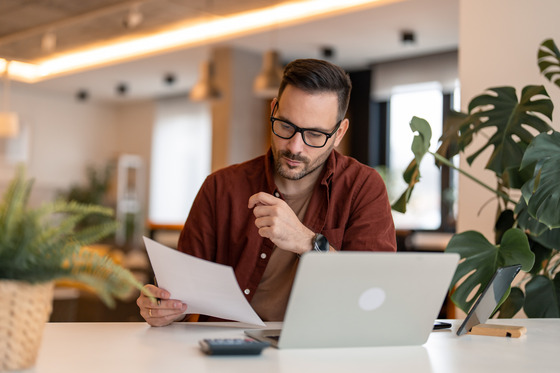 Adult male reading documents in paper and using his laptop.