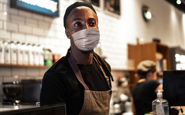 Young man wearing a COVID mask and working behind a counter at a coffee shop. 