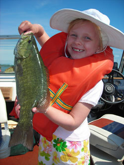 A young girl in a wide brimmed white hat and orange life preserver holding up a smallmouth bass