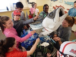 Students gather around a sculpture of a striped bass made entirely of plastic debris found on coastal shores.