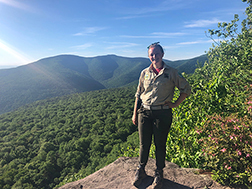 A NY NJ trail conference steward at the top of giant ledge with a scenic view of the catskill mountains in the background