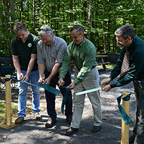 A group of DEC operations staff cutting a green ribbon across an accessible campsite that is now open at Nick's Lake campground